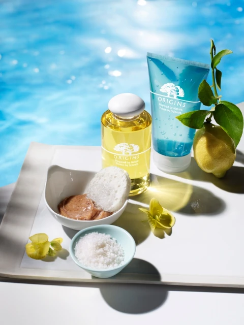 Beauty still life: Origins skincare products displayed on a white surface with lemon, sea salt, and a swimming pool background.