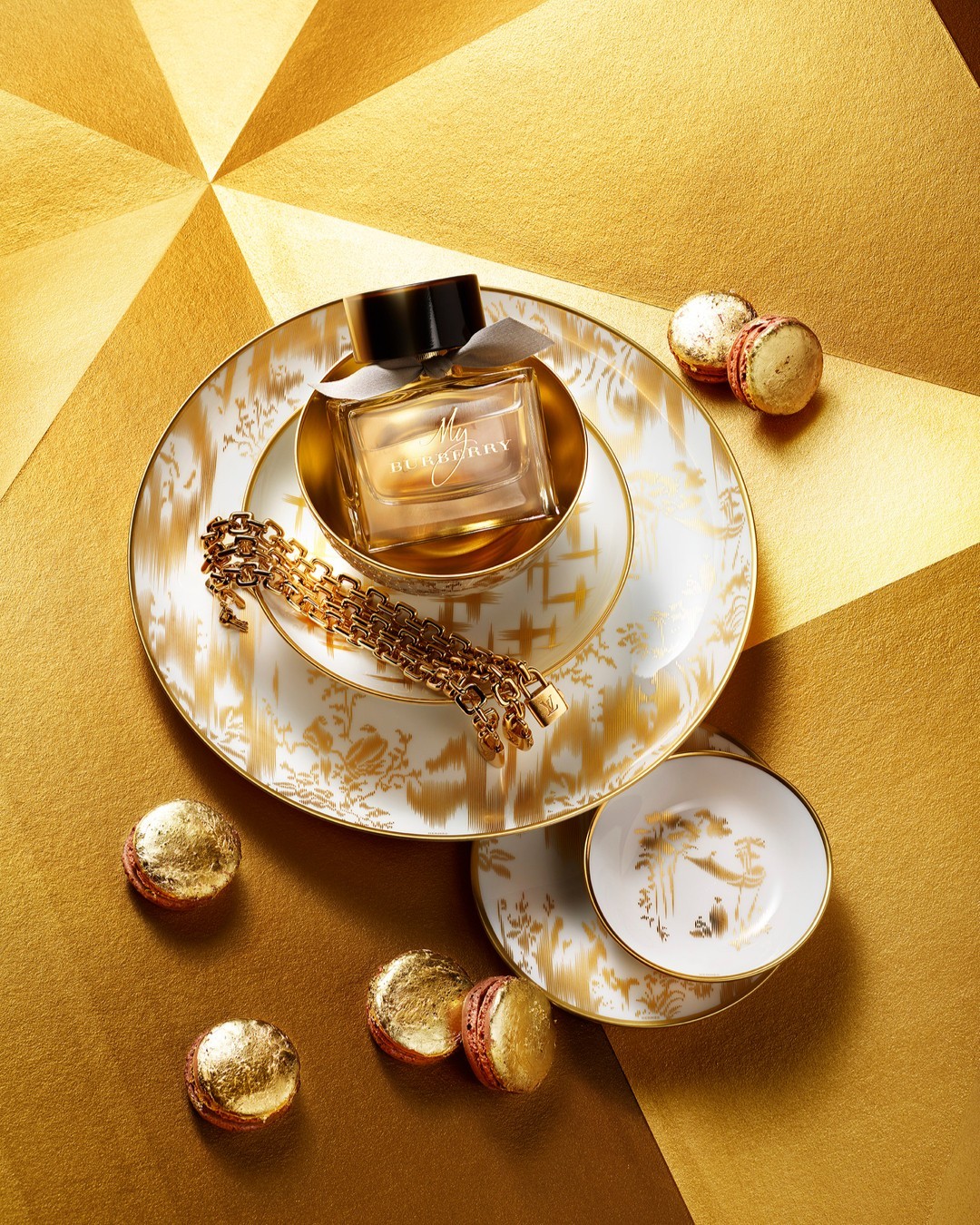 A bottle of Burberry Her Elixir de Parfum sits on a porcelain plate alongside macarons and a gold chain on a geometric gold surface.