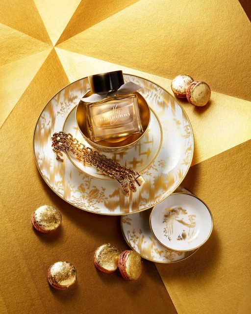 A bottle of Burberry Her Elixir de Parfum sits on a porcelain plate alongside macarons and a gold chain on a geometric gold surface.