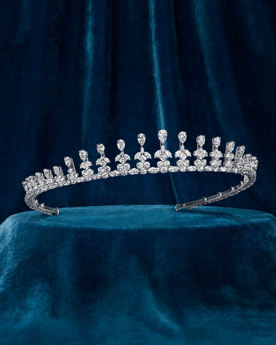 A silver tiara with many crystals rests on a blue velvet surface, displayed in a studio with soft lighting.