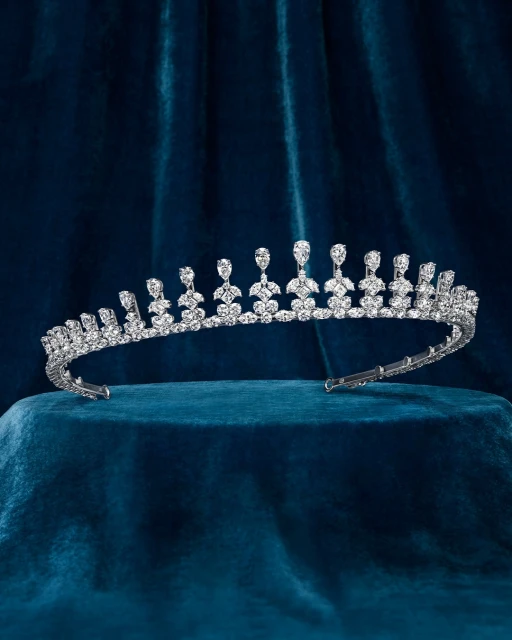 A silver tiara with many crystals rests on a blue velvet surface, displayed in a studio with soft lighting.