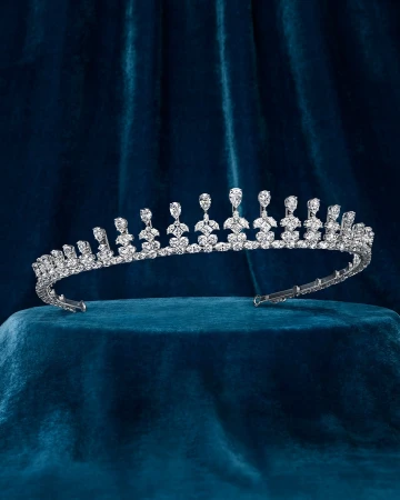 A silver tiara with many crystals rests on a blue velvet surface, displayed in a studio with soft lighting.
