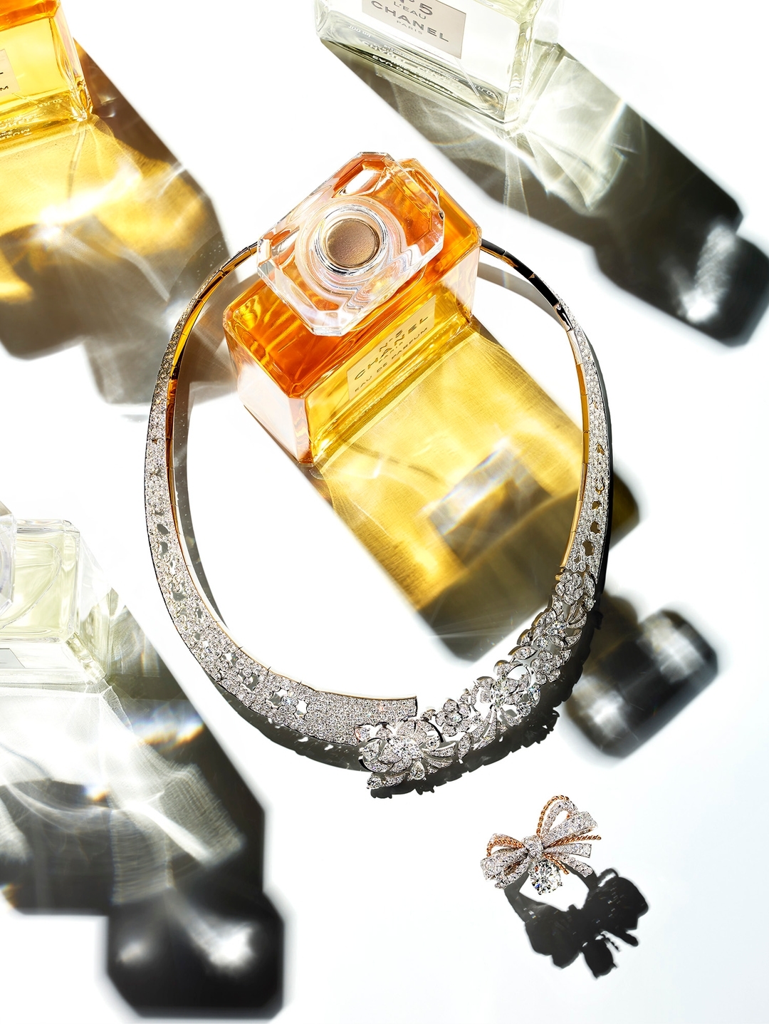 Still life of jewelry and Chanel perfume bottles in studio light featuring a crystal necklace, crystal bow, and glass perfume bottles.