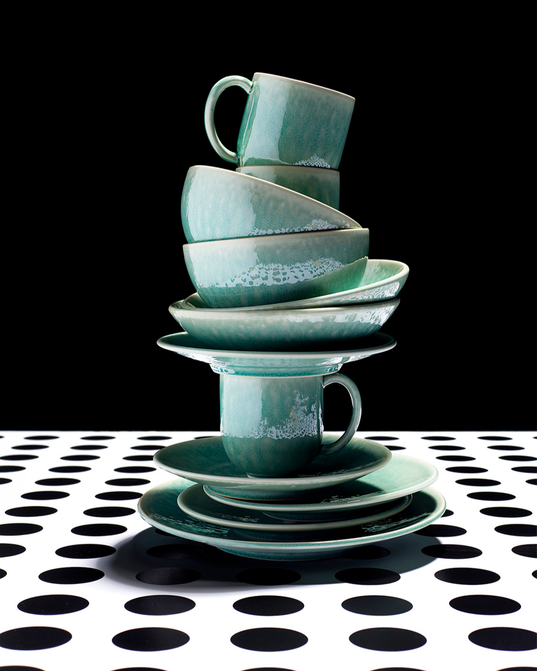 Stack of teal ceramic tableware including plates, bowls, and cups on a polka dot tablecloth against a black background.
