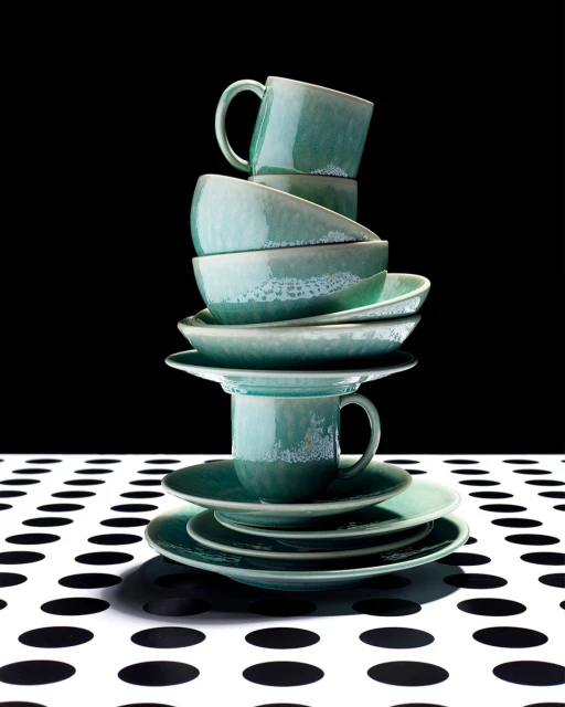 Stack of teal ceramic tableware including plates, bowls, and cups on a polka dot tablecloth against a black background.