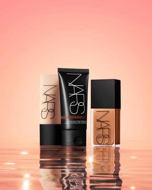 Three NARS foundation products in varying shades are displayed against a blurred coral background with a reflective surface. 