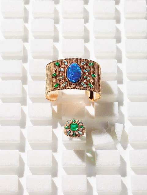 Close-up of an opulent gemstone-encrusted gold bracelet and ring set against a stark white tile background with directional lighting.