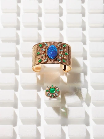 Close-up of an opulent gemstone-encrusted gold bracelet and ring set against a stark white tile background with directional lighting.