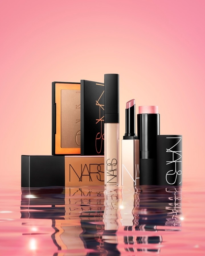 NARS makeup products including bronzer, concealer, lipstick, and blush are displayed on a reflective surface against a pink background.