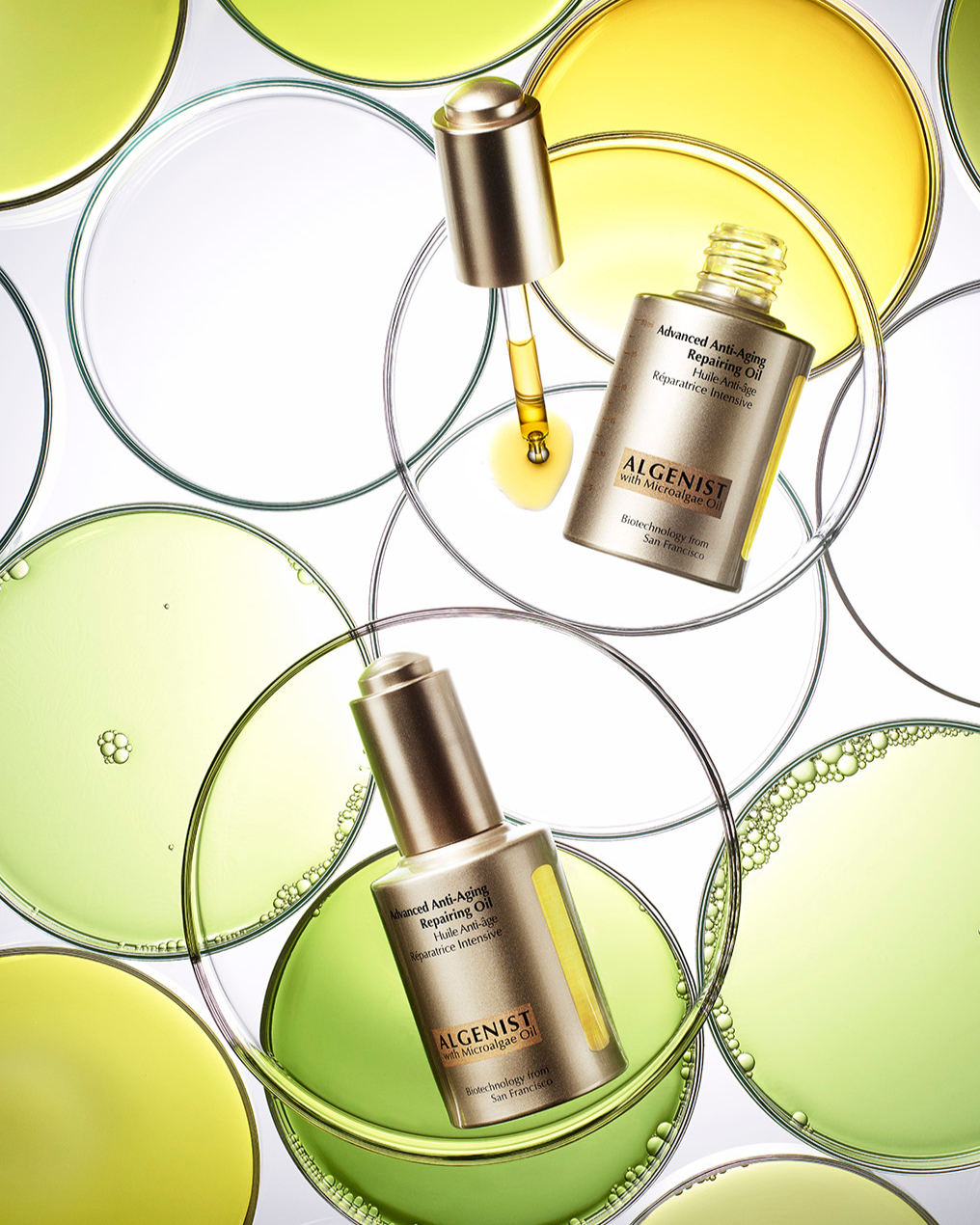 Cosmetic product shot featuring Algenist Advanced Anti-Aging Repairing Oil bottles with glass droppers, surrounded by petri dishes filled with liquids.