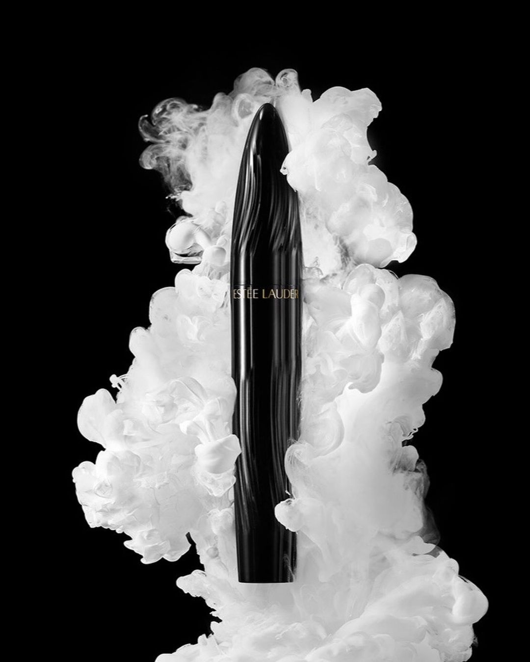 Elegant Estee Lauder mascara tube shown against a backdrop of swirling white smoke on a stark black background in a beauty product still life.