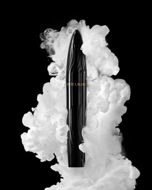 Elegant Estee Lauder mascara tube shown against a backdrop of swirling white smoke on a stark black background in a beauty product still life.