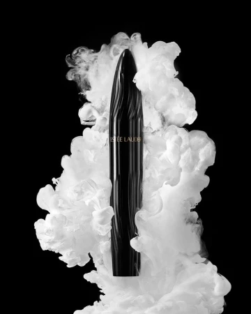 Elegant Estee Lauder mascara tube shown against a backdrop of swirling white smoke on a stark black background in a beauty product still life.