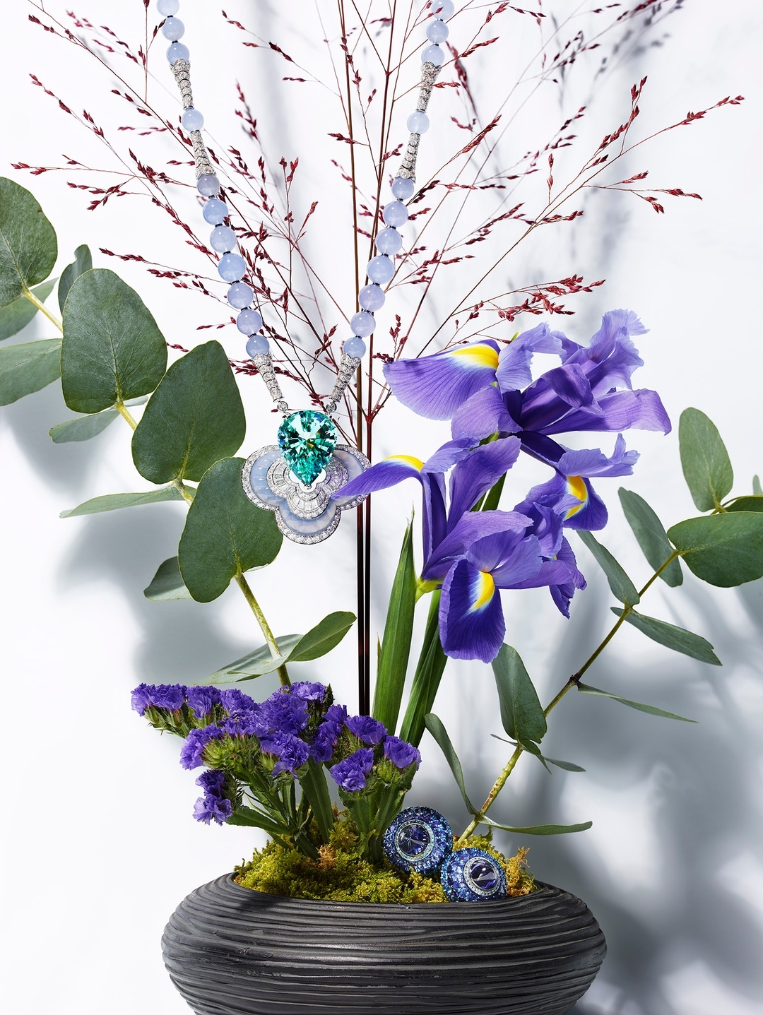 Elegant jewelry necklace featuring blue gemstones and a green pendant, displayed amidst purple iris, eucalyptus, and delicate branches in a vase.