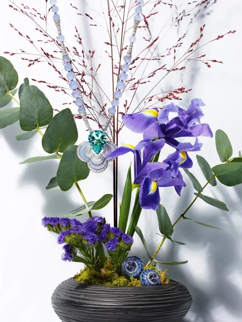 Elegant jewelry necklace featuring blue gemstones and a green pendant, displayed amidst purple iris, eucalyptus, and delicate branches in a vase.