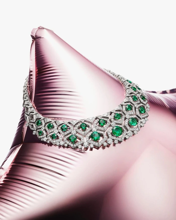 A diamond and emerald necklace draped over a shiny pink metallic balloon against a white background.