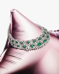 A diamond and emerald necklace draped over a shiny pink metallic balloon against a white background.