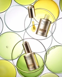 Overhead product photography of Algenist serum bottles and dropper with yellow oil inside glass petri dishes.