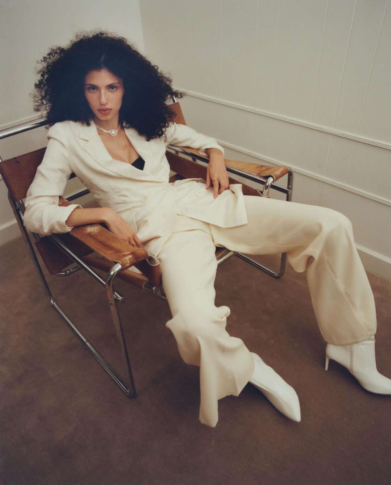 Woman with voluminous curly hair wearing a cream pantsuit and white boots seated in a chrome armchair