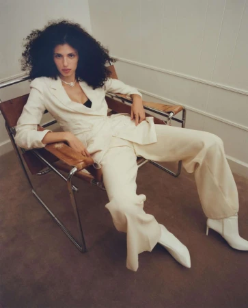 Woman with voluminous curly hair wearing a cream pantsuit and white boots seated in a chrome armchair