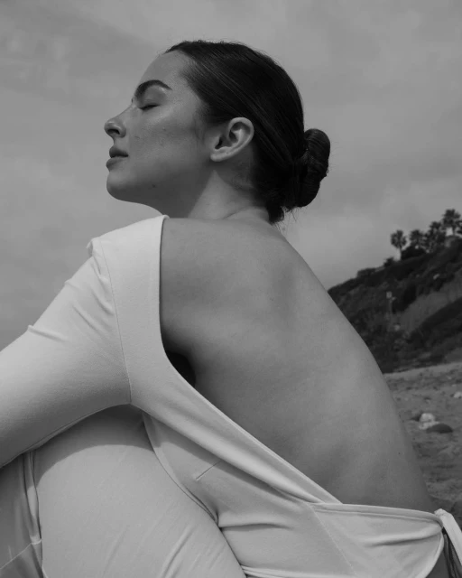 Adult woman in a light-colored outfit, captured in a black and white outdoor portrait, eyes closed, seated with a beach and sky backdrop.