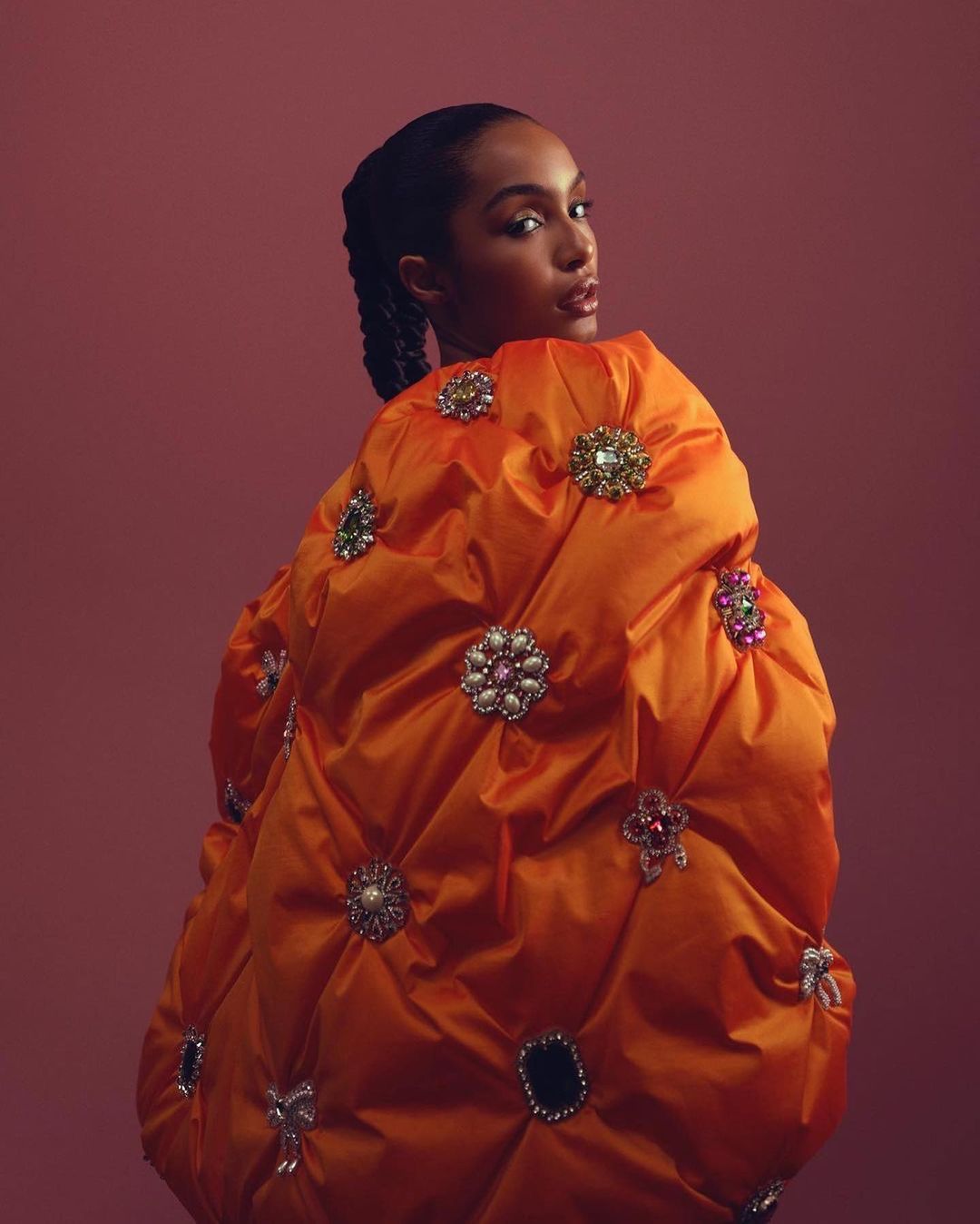 Elegant Black female model with braided hair wears orange statement jacket adorned with decorative brooches in a studio fashion shot.