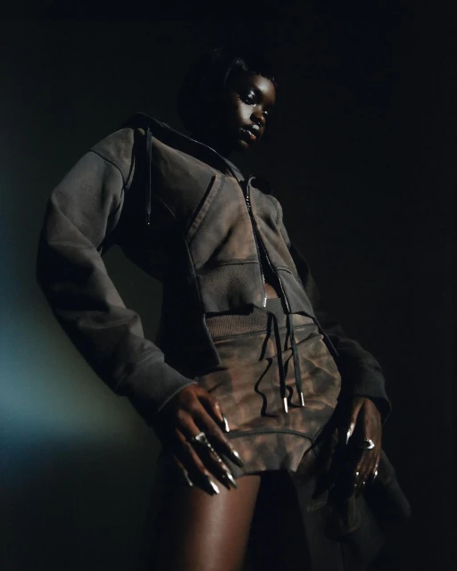 Woman in a grey tie-dye hoodie and shorts poses in a studio setting, illuminated by a spotlight against a dark background.