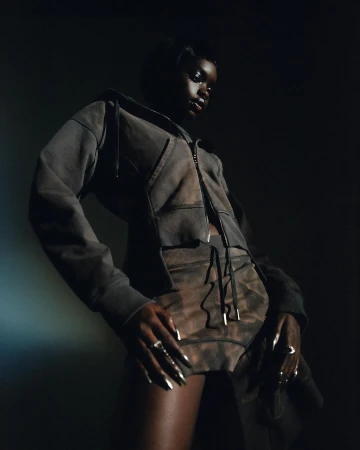 Woman in a grey tie-dye hoodie and shorts poses in a studio setting, illuminated by a spotlight against a dark background.