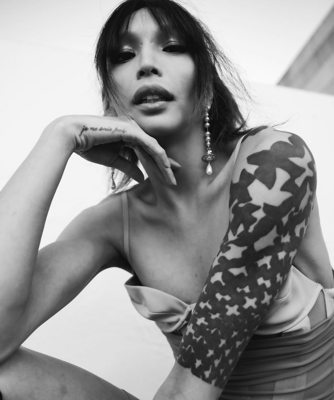 Black and white portrait of a person with dark hair and tattoos posing with their hand near their face, wearing earrings.
