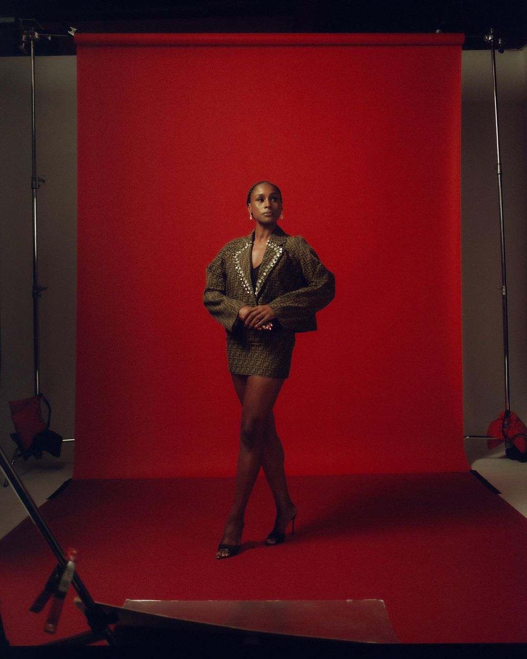 A stylish adult woman in a blazer and mini skirt poses against a red backdrop in a photo studio setting with visible lighting equipment.