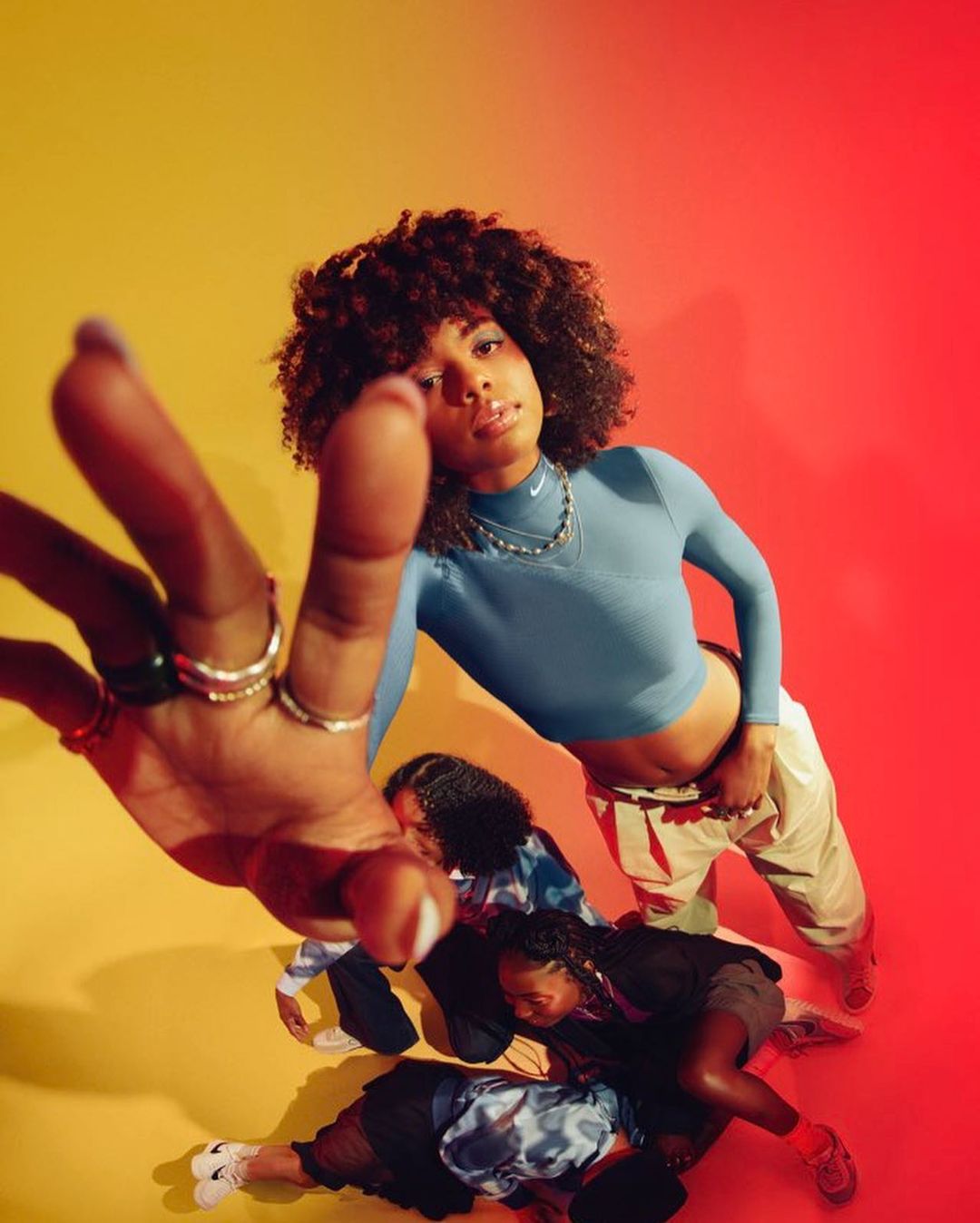 Young woman with Afro in athletic top extends hand to camera with three other people in the background in a studio with yellow and red lighting.