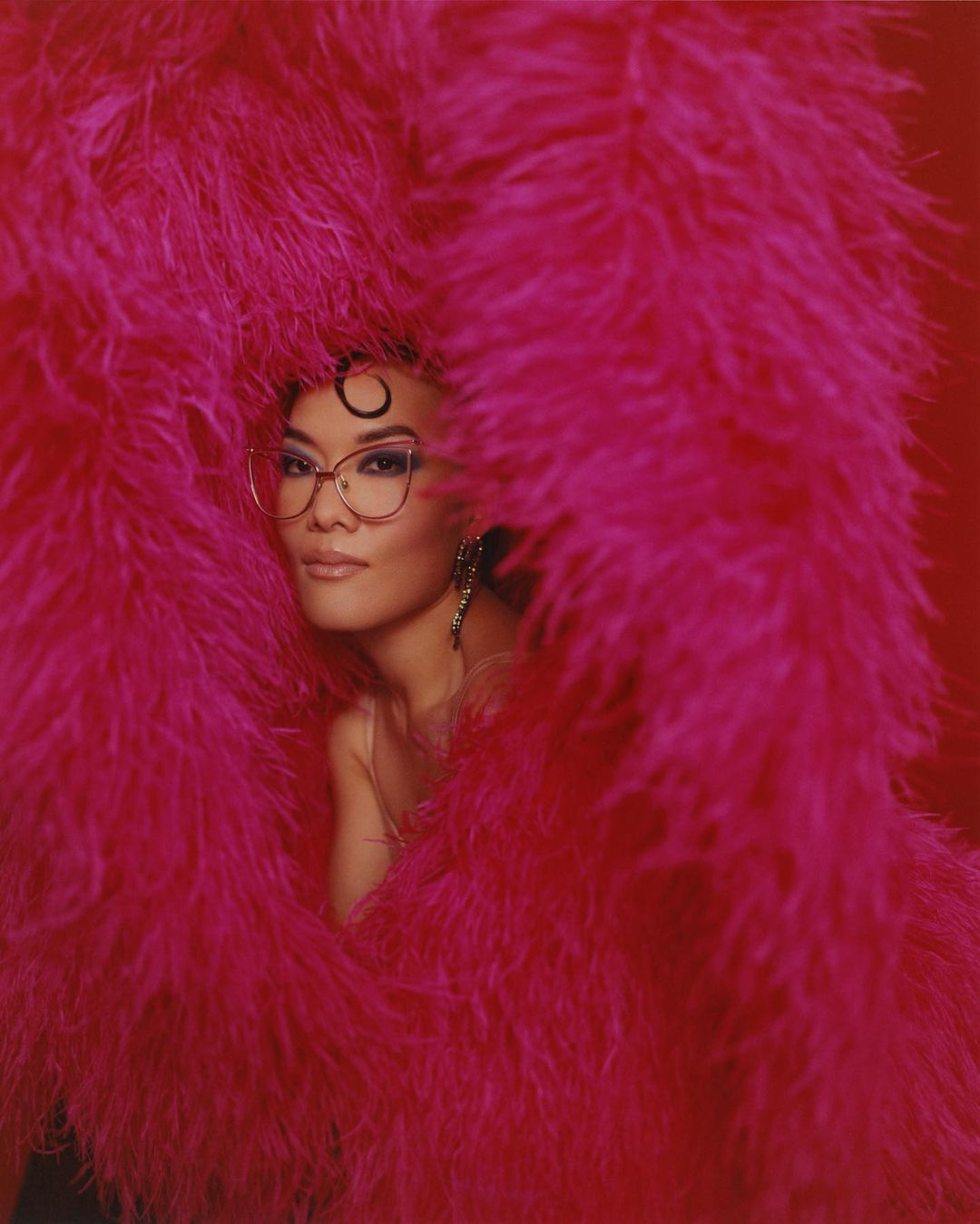 Portrait of Asian woman with short dark hair, glasses, earrings, and fuchsia feather boa against a red background in a photo studio.