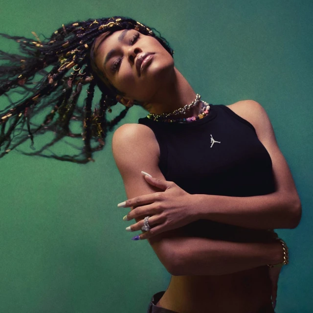 Woman with locs in a black tank top poses against a green background, showcasing jewelry and stylish nails. Fashion and beauty visual.