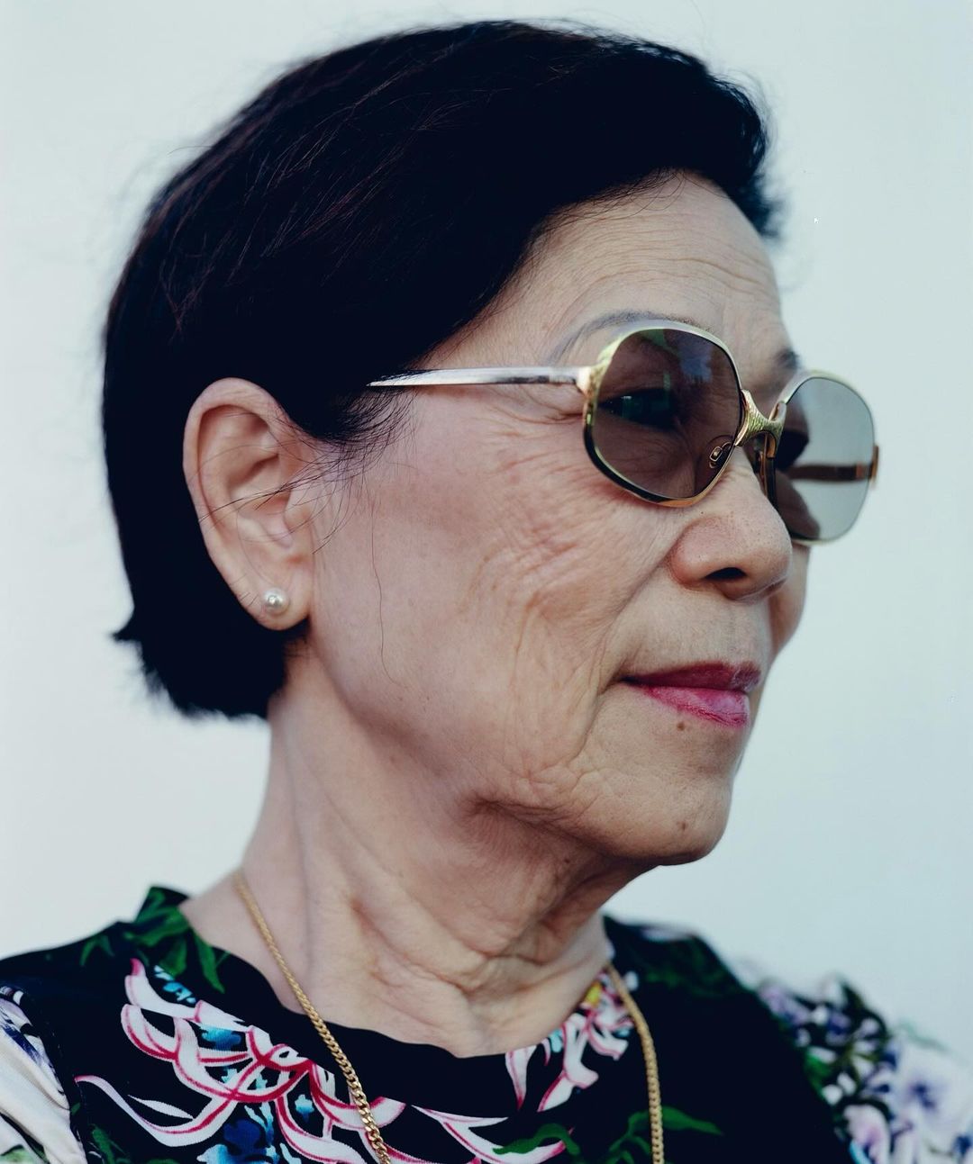 Close-up portrait of an older woman with short dark hair wearing tinted sunglasses, a floral shirt, and a gold chain necklace. 