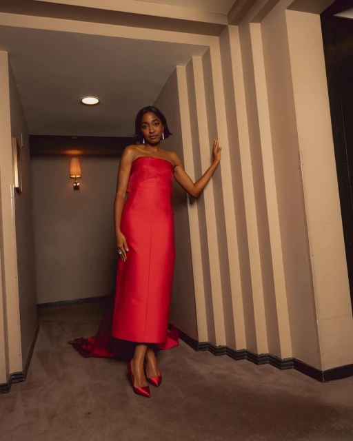Elegant adult woman in a red strapless gown and matching shoes poses in an indoor hallway with striped wall paneling.