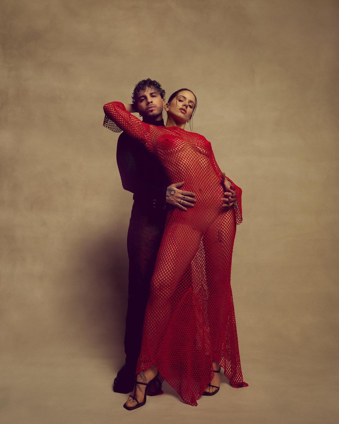 Sensual studio portrait of a couple: a tattooed man in black embraces a woman in a red fishnet dress and black high heels against beige backdrop.