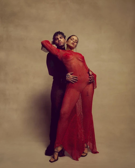 Sensual studio portrait of a couple: a tattooed man in black embraces a woman in a red fishnet dress and black high heels against beige backdrop.