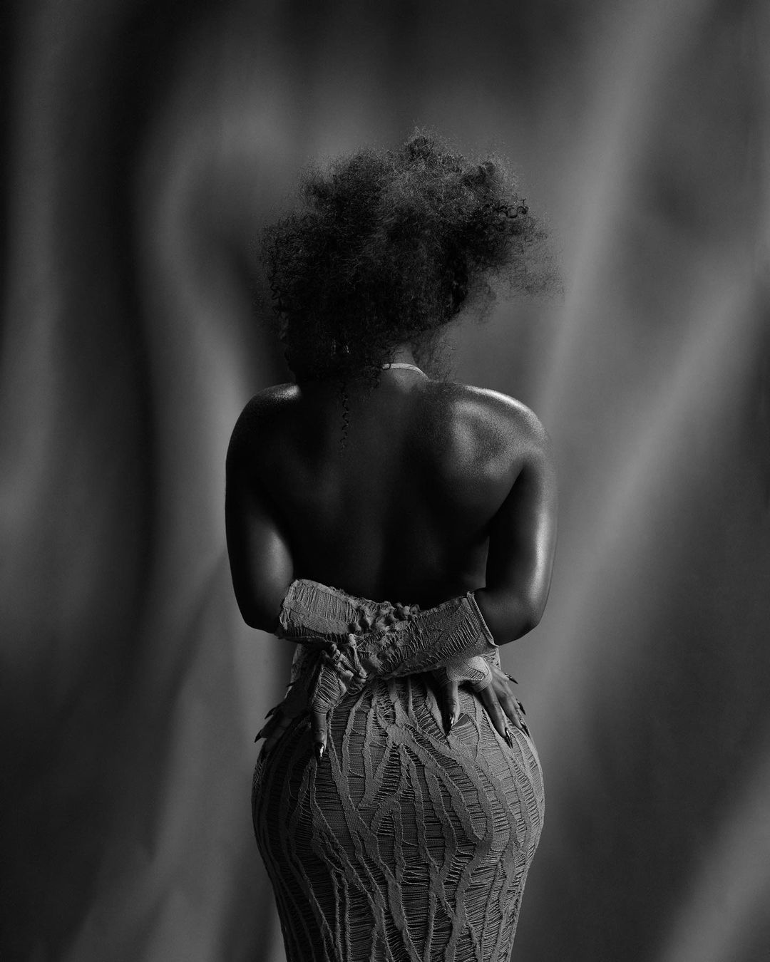 Monochrome portrait of Black woman with afro, facing away from camera, wearing patterned dress, hands on hips against a blurred backdrop.