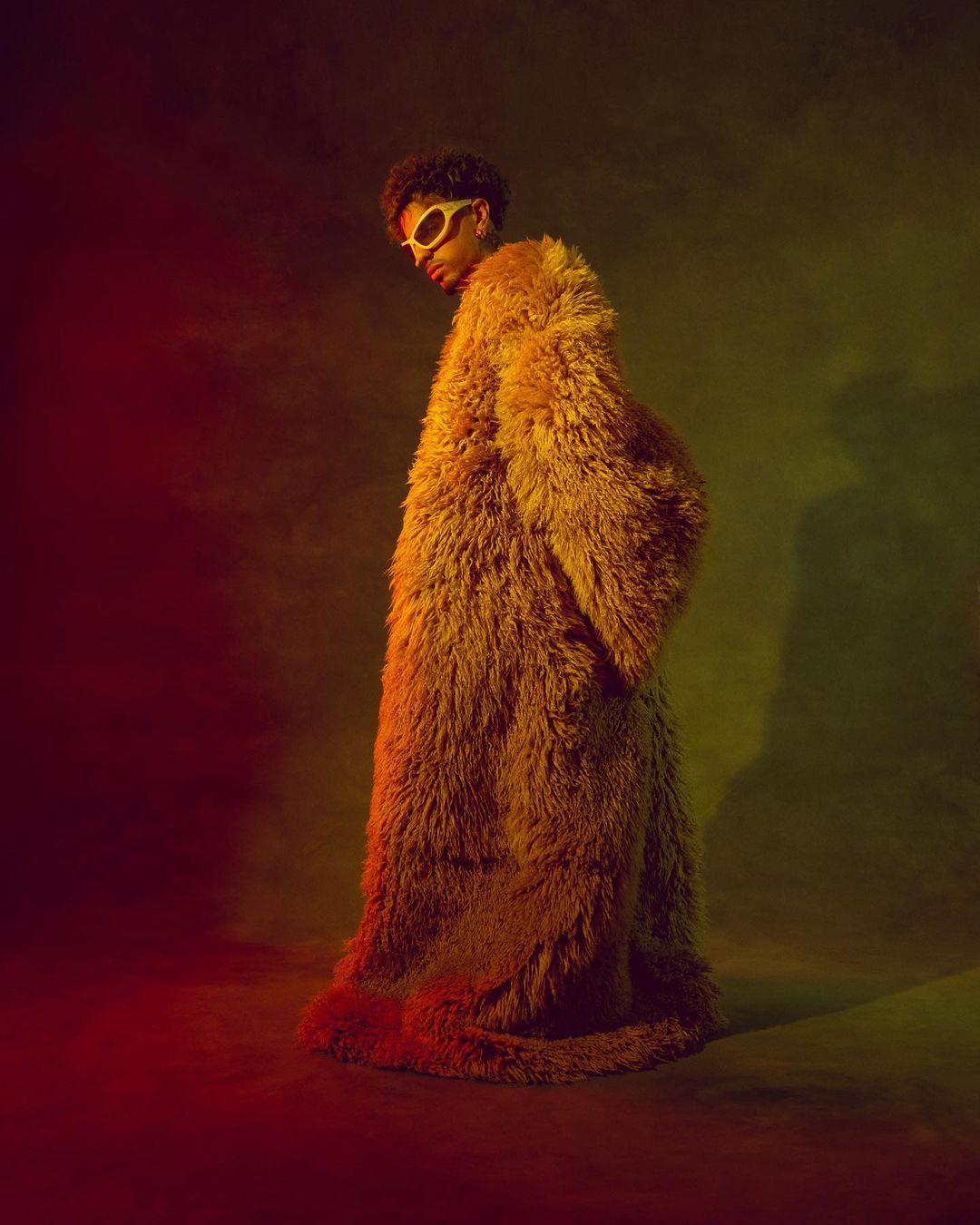 A person with afro hair and sunglasses poses in a yellow, faux fur coat against a blurred background with red and green lighting.