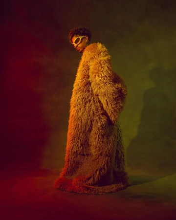 A person with afro hair and sunglasses poses in a yellow, faux fur coat against a blurred background with red and green lighting.