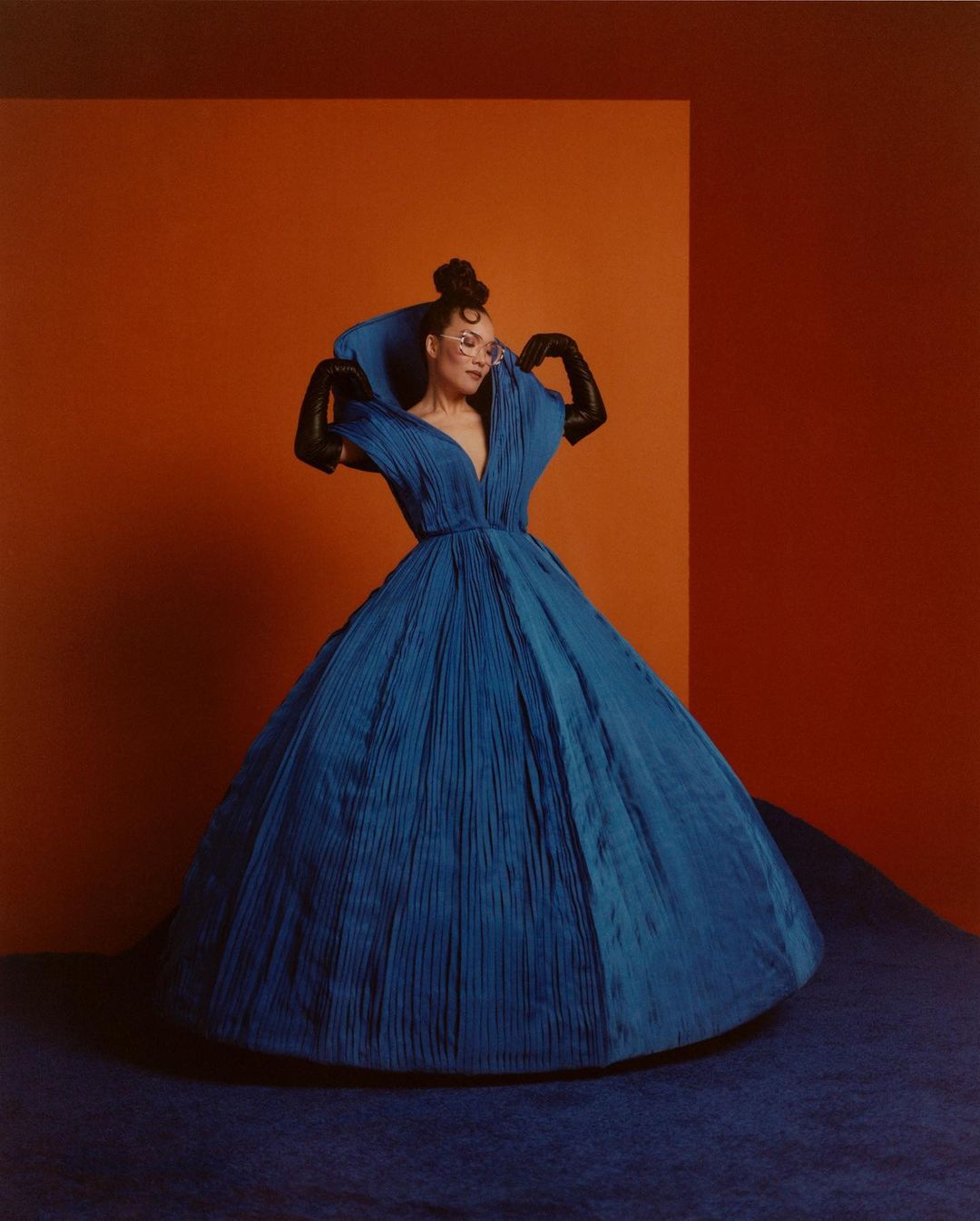 Woman in a vibrant blue, pleated ballgown poses against a contrasting orange and red backdrop, creating a striking fashion statement.