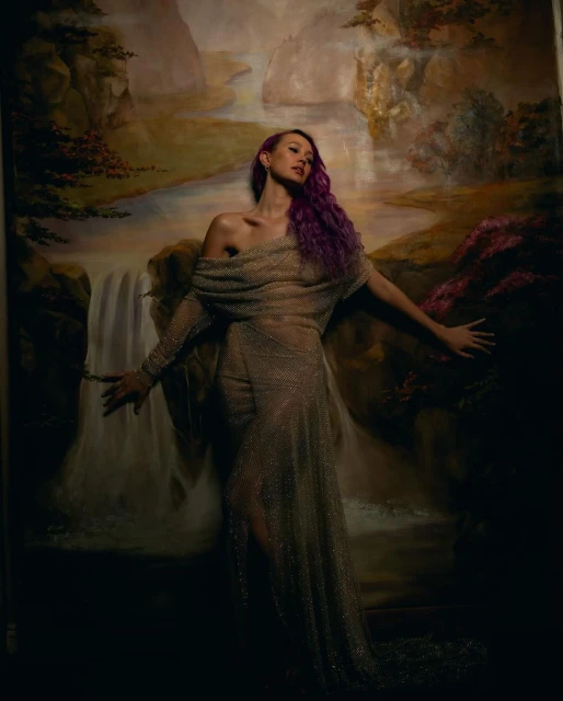 Woman in sheer gown poses against a painted backdrop featuring waterfall and foliage; she has violet hair and light skin.