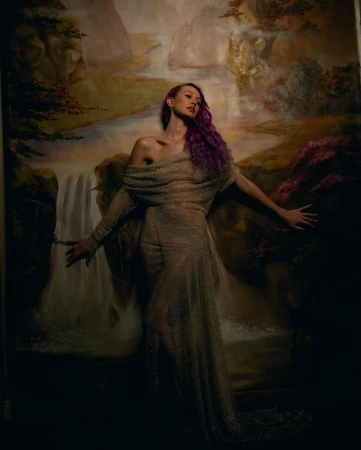 Woman in sheer gown poses against a painted backdrop featuring waterfall and foliage; she has violet hair and light skin.