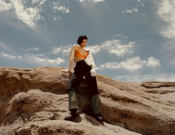 Model wearing orange bralette and dark trousers perched on a rocky outcrop against a cloudy blue sky.