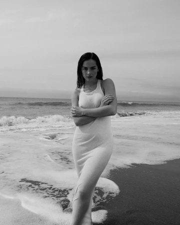 Woman in a long white dress standing on a sandy beach with ocean waves in the background, captured in black and white.