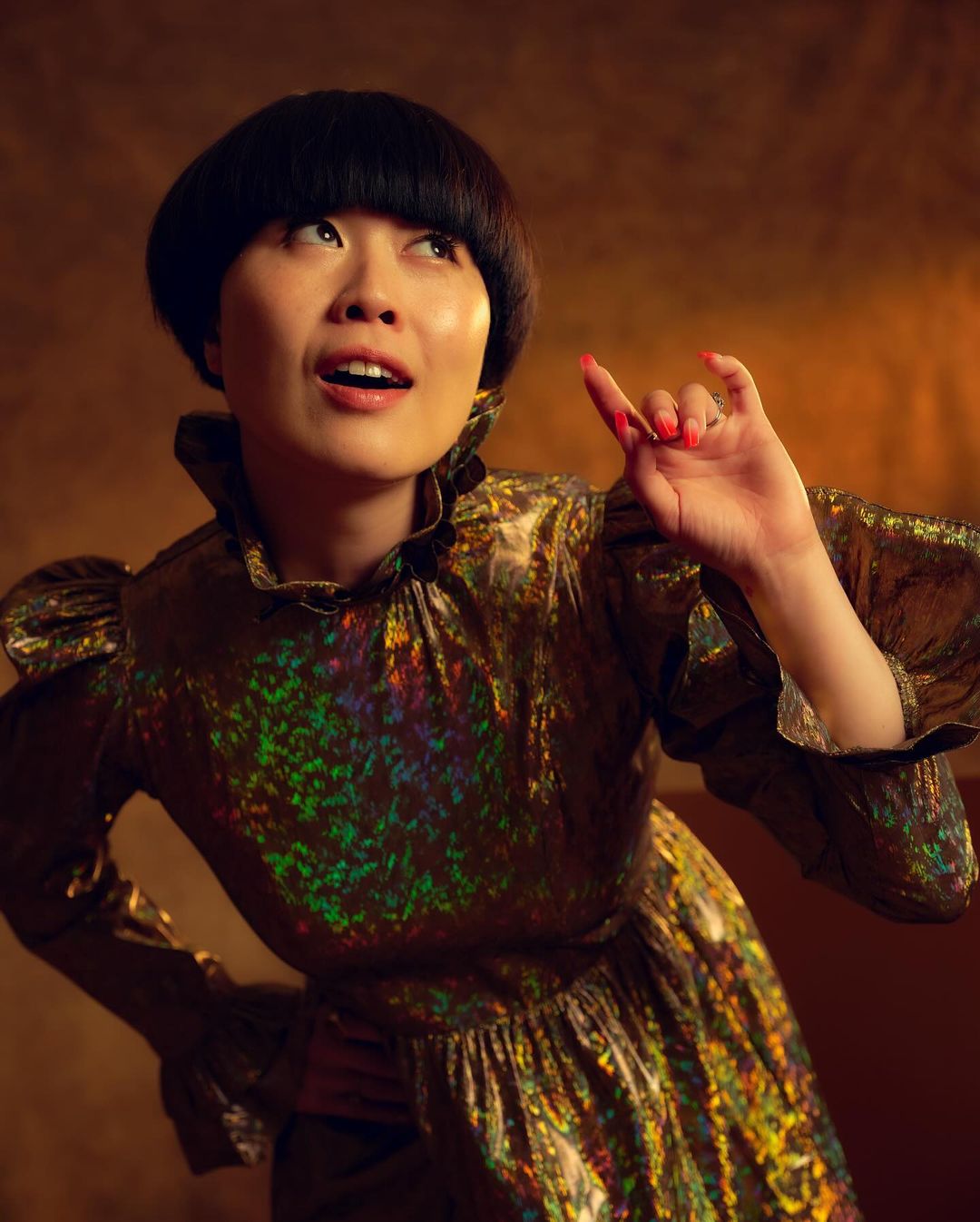 Portrait of a young Asian woman in a gold dress, looking up with a slight smile, set against a textured amber and gold background.