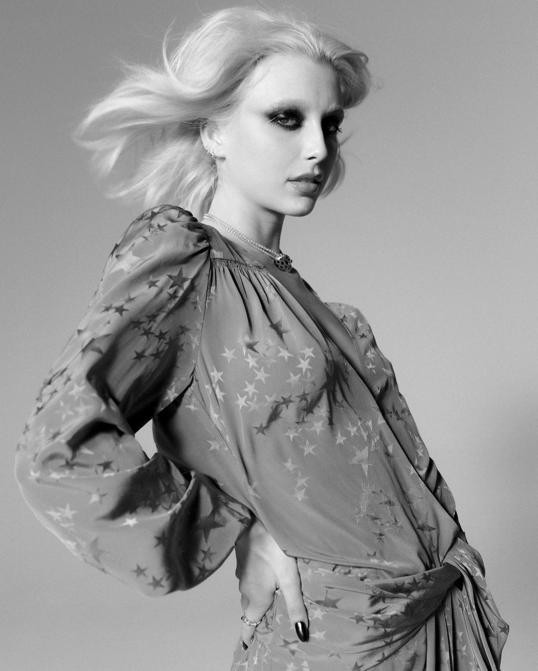 Monochrome portrait of a model with windswept blond hair wearing a star-patterned dress against a neutral backdrop in a fashion-style image.