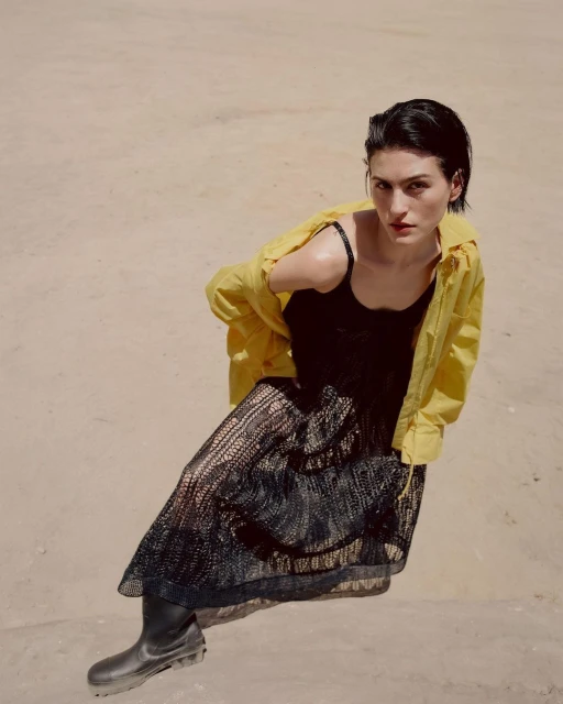 A woman with short dark hair, in a black dress and yellow jacket, poses in an outdoor shot with sandy ground as the backdrop.