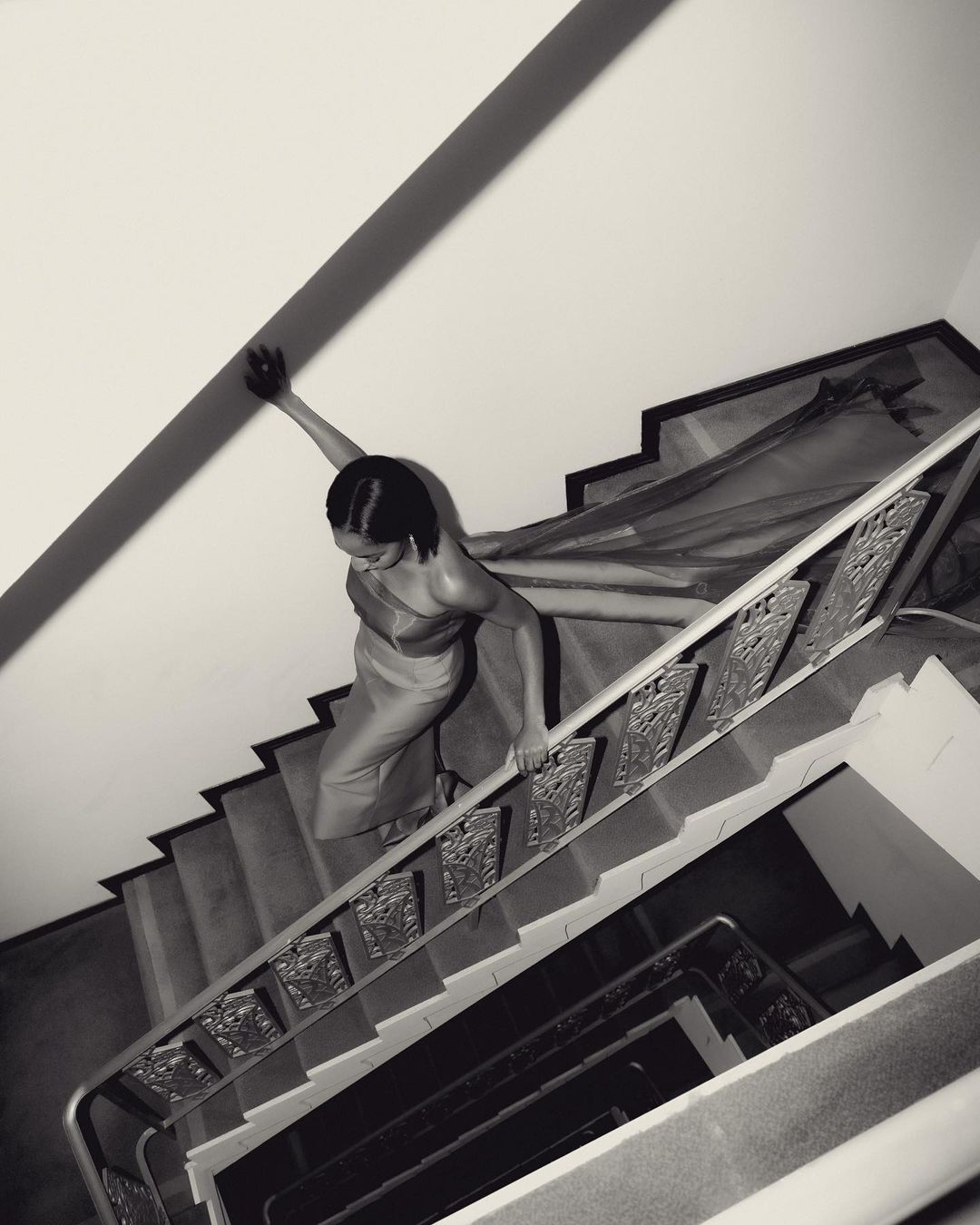 Woman in dress climbs an indoor staircase with ornate metal railing. Black and white photography with high-angle perspective.