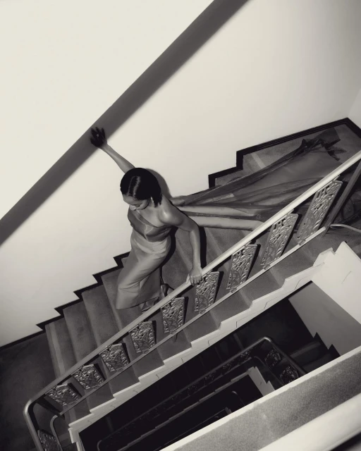 Woman in dress climbs an indoor staircase with ornate metal railing. Black and white photography with high-angle perspective.